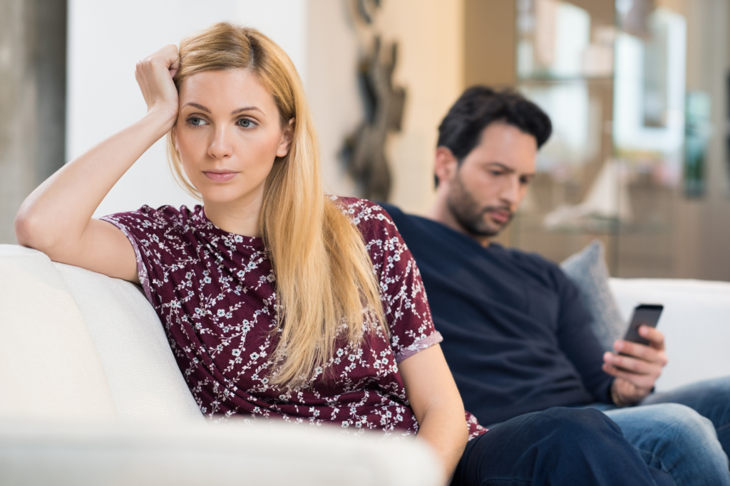 A couple sitting separately on a sofa during an argument, with the husband looking at his phone while the wife faces away, appearing upset and distant