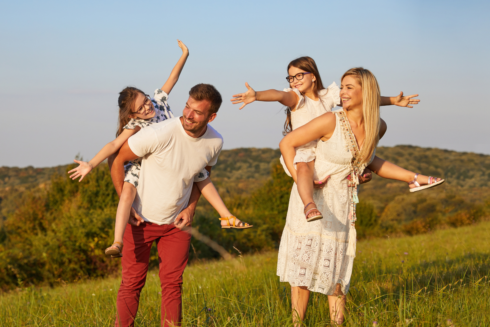 Parents playing with their daughters outdoors A mother carrying her daughter on her back and a father lifting their other daughter while playing outside on a sunny day