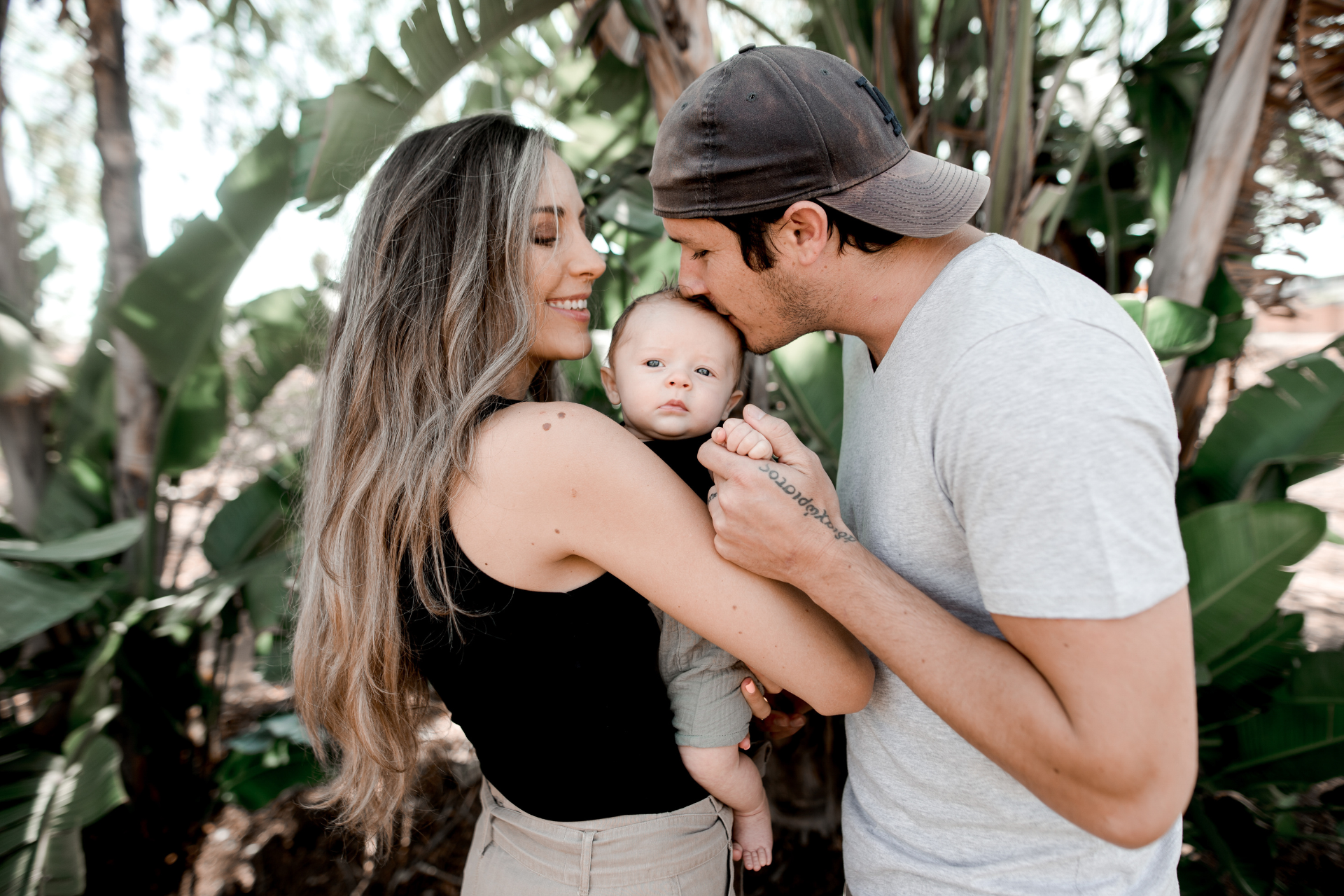 A mother holding her baby up while the father kisses the baby, standing outside with a tree visible in the background