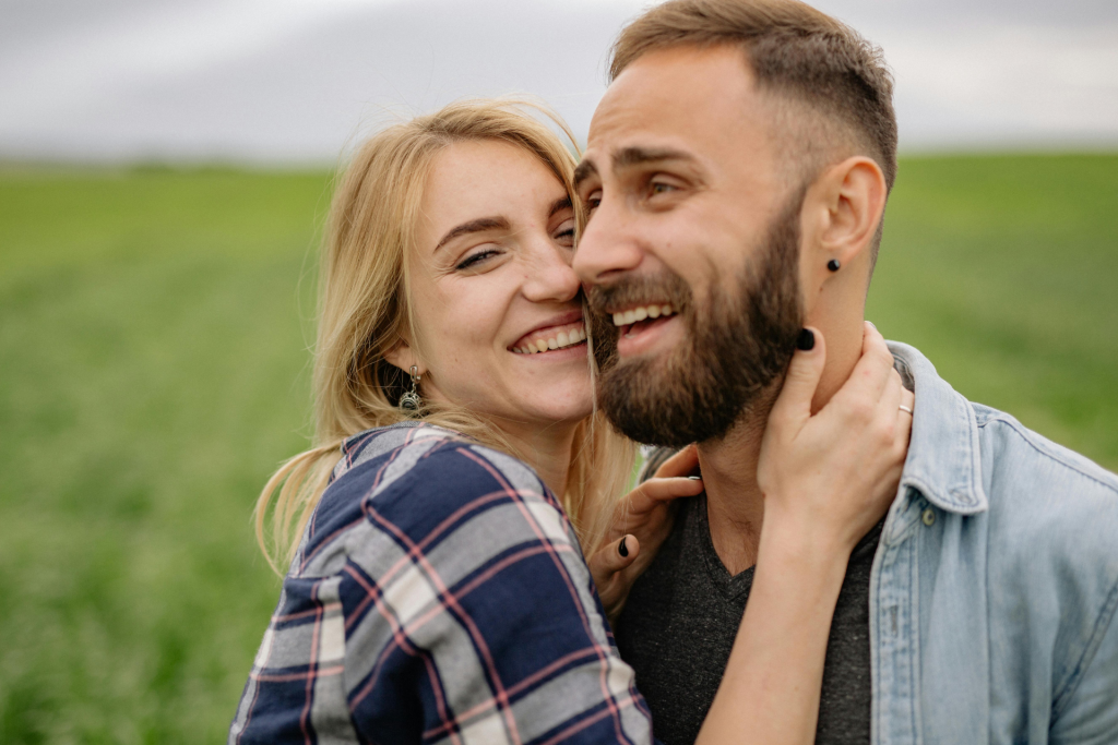 A close-up of a smiling couple outdoors, with the woman holding the man by the neck as they laugh and lean in for a kiss, green grass blurred in the background