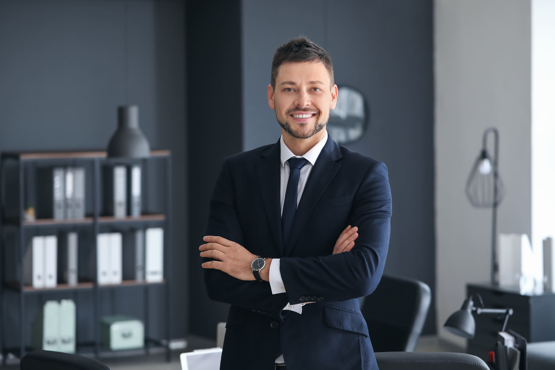 A well-dressed man in an elegant suit standing in an office with his arms crossed, conveying confidence and professionalism