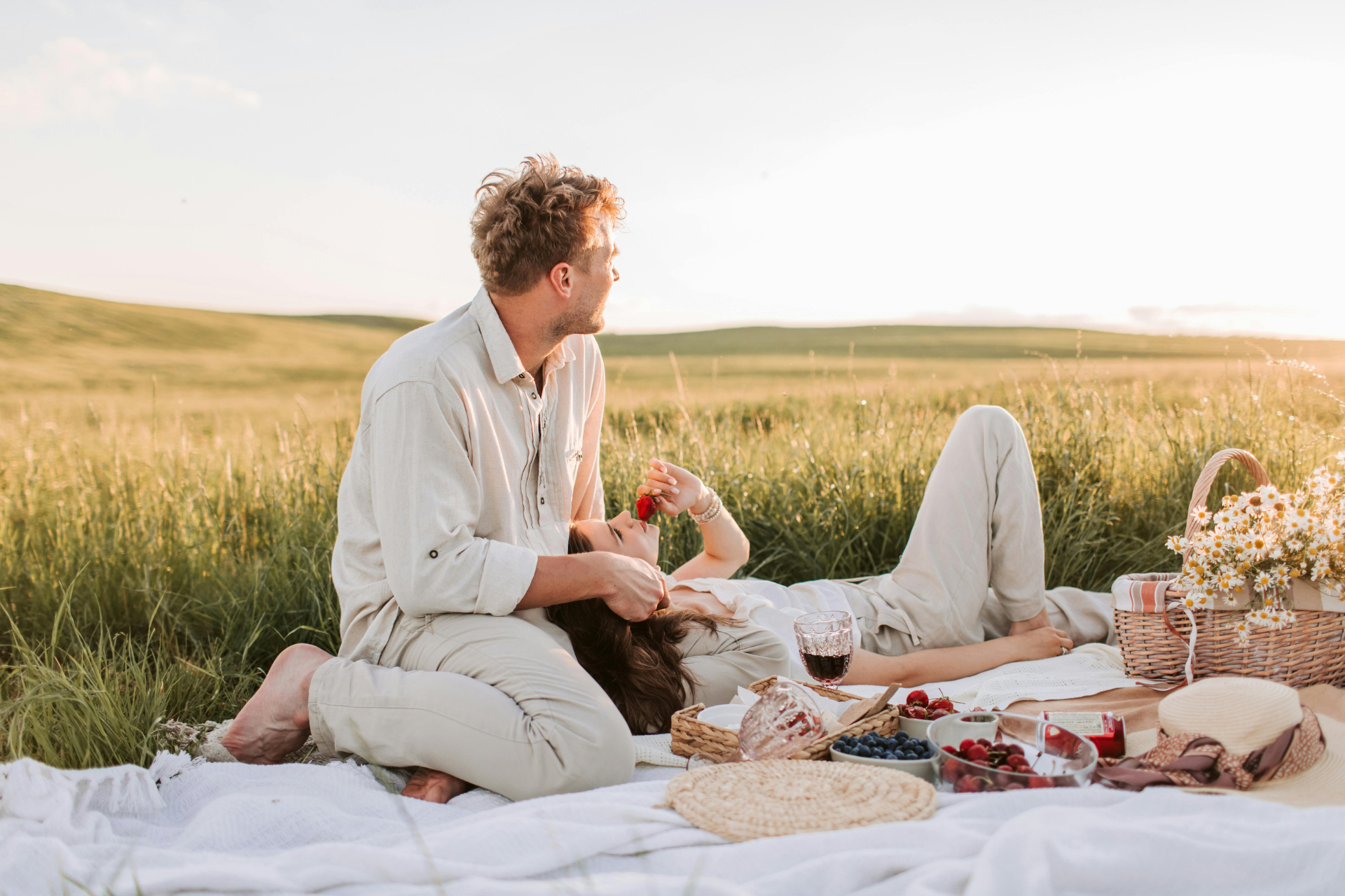 A couple enjoying a sunset picnic, with the woman resting on the man’s legs as they look toward the horizon, sharing fruit and wine together