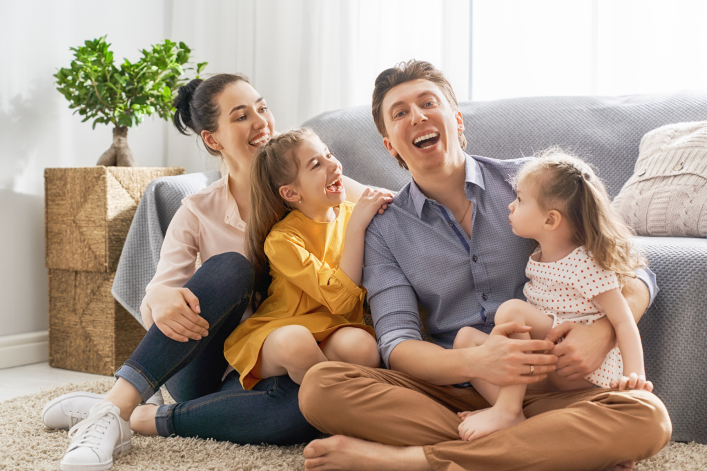 A happy family with two daughters and their parents sitting on the floor in front of a sofa, smiling together in their living room