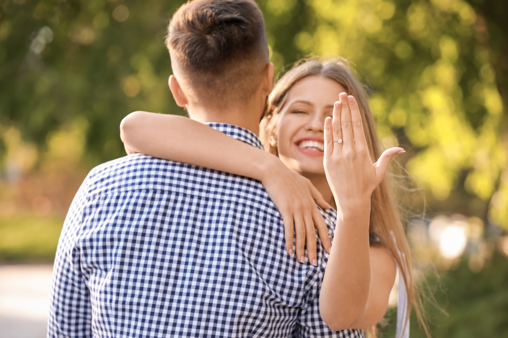 Woman showing her engagement ring while hugging her partner A smiling woman holding up her engagement ring to the camera as she embraces her partner on a sunny day, with the man seen from behind