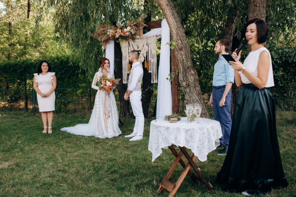 A couple standing together during their wedding ceremony, listening closely as a speaker delivers a reading in front of their guests