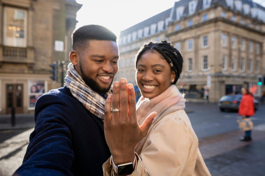 A couple hugging with a city in the background as the woman shows her engagement ring toward the camera, highlighting the proposal moment