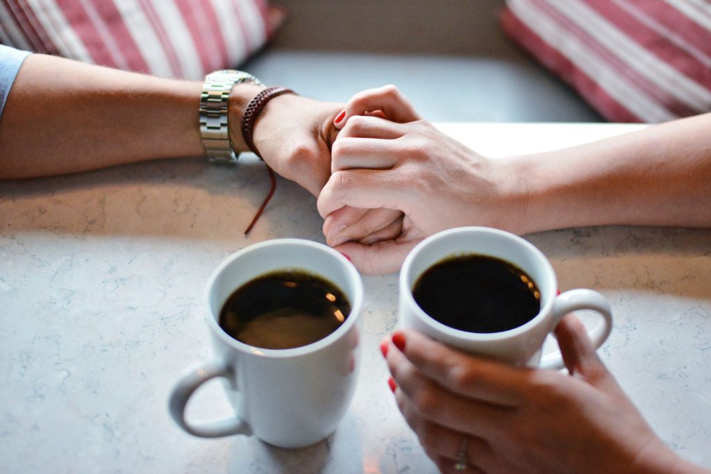 A close-up of a couple holding hands across a table with a cup of coffee, showing only their hands in a warm, intimate setting