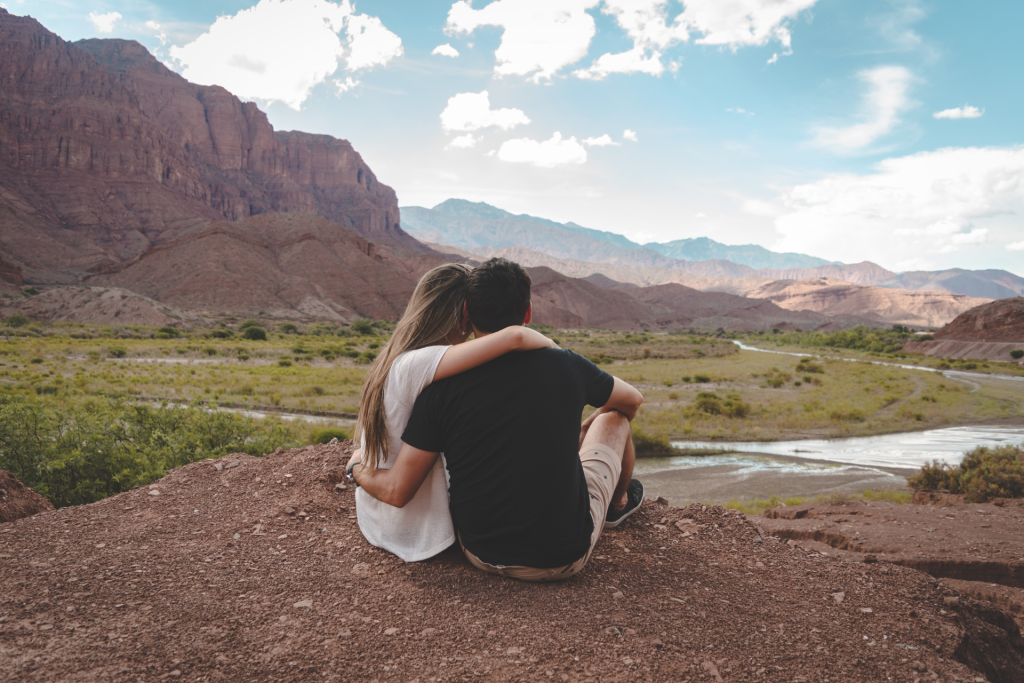 A couple sitting side by side with their backs to the camera, looking out at a scenic view of mountains and a river in the distance