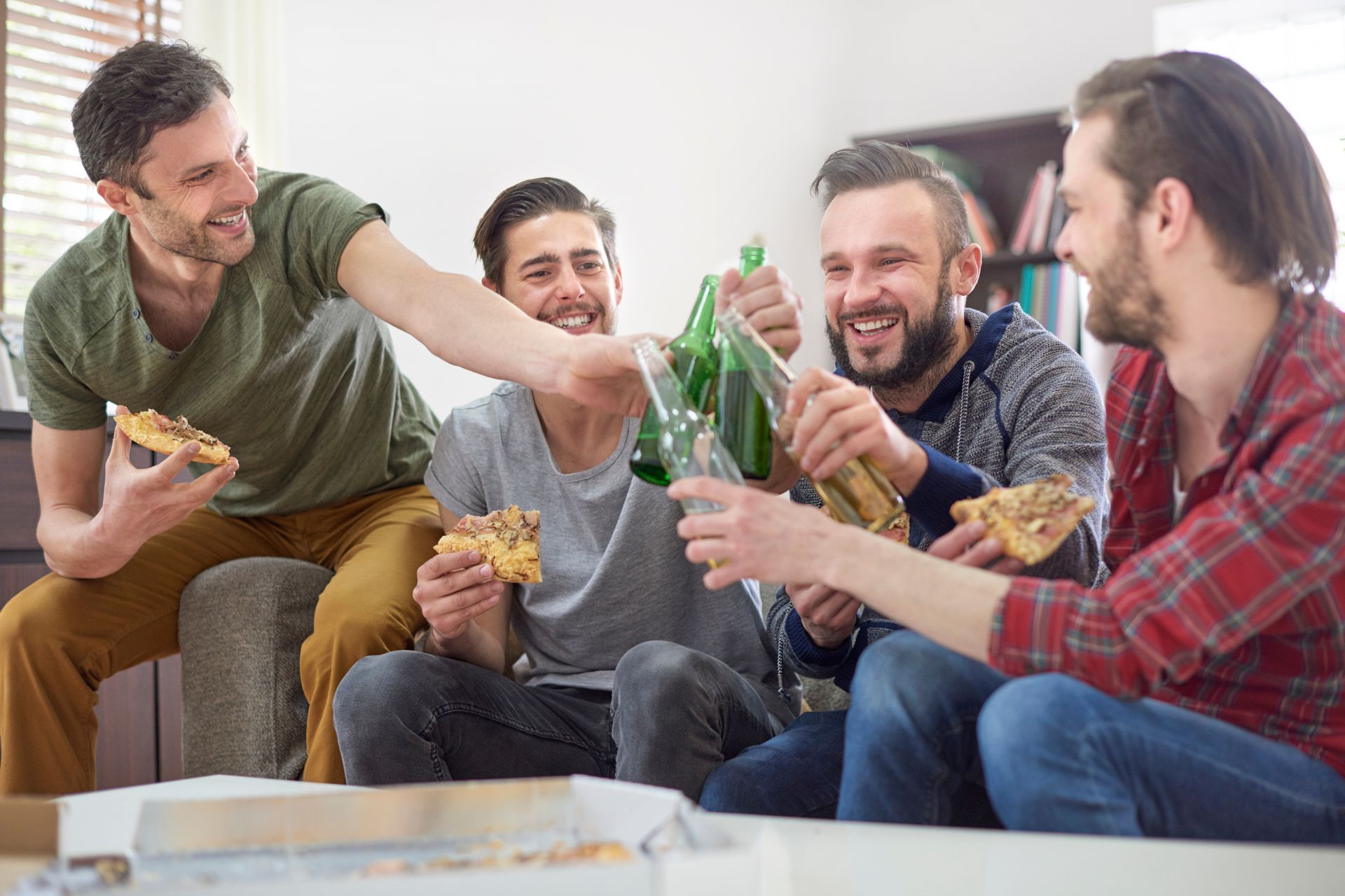 Four friends raising their beers in a toast while sharing pizza together and enjoying a relaxed gathering