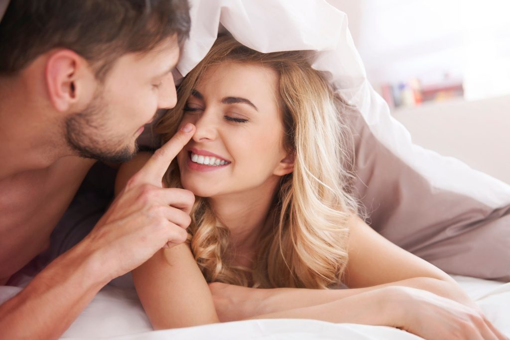 A couple wrapped in a blanket, where the man gently touches the woman’s nose in a playful gesture as she smiles with her eyes closed