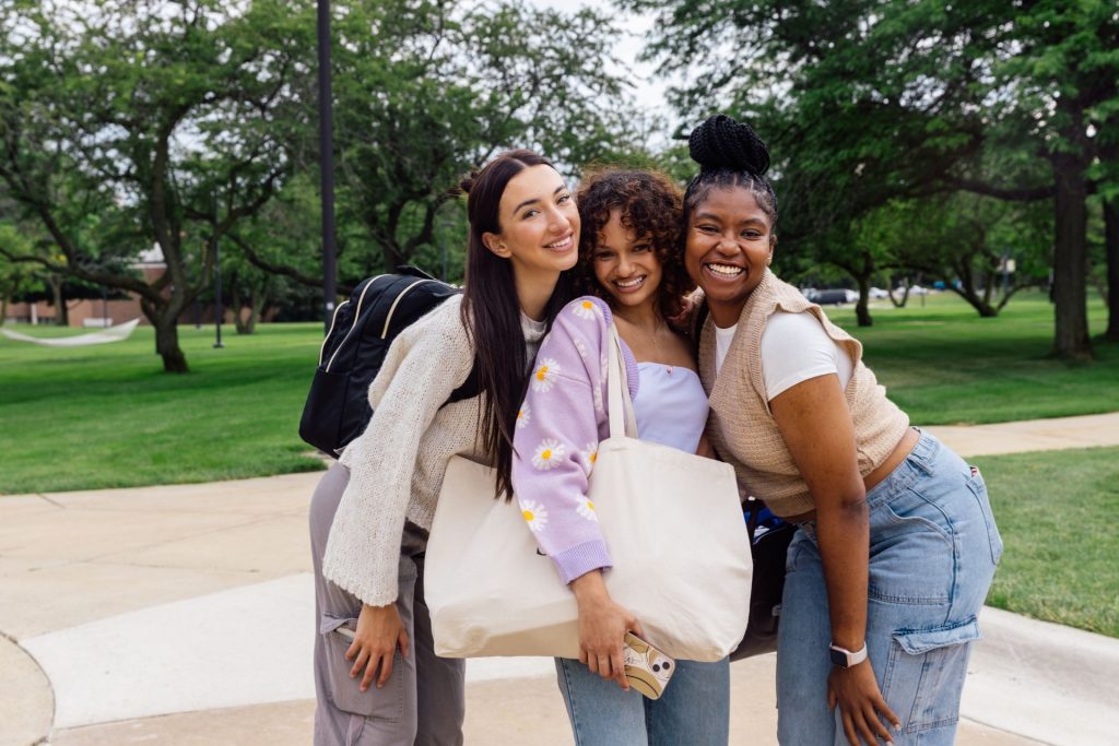 Three friends smiling and posing for a photo in what appears to be a park or university campus with greenery in the background