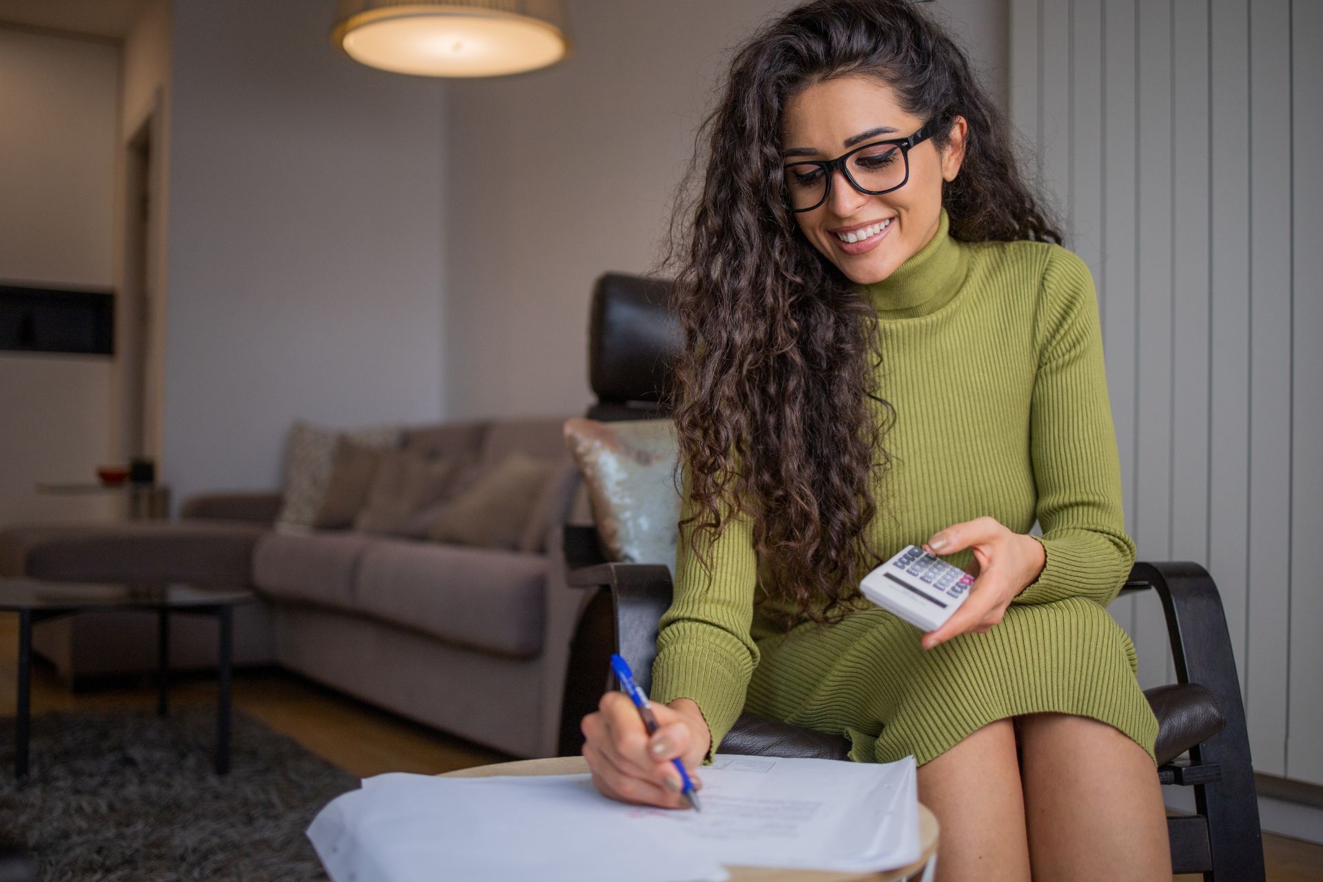 A woman holding a calculator while writing on papers at a table, smiling calmly as she reviews financial documents