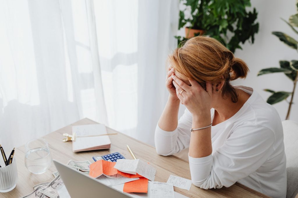 A woman sitting at a table with financial papers and documents, holding her head in concern while reviewing her finances