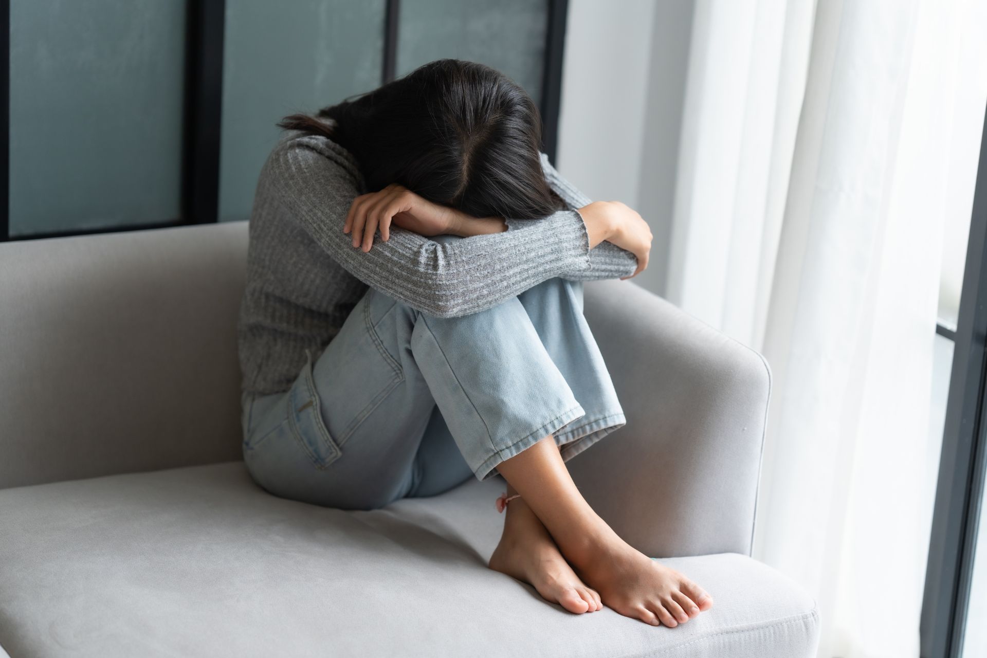 A woman sitting on a sofa hugging her legs, appearing worried and overwhelmed based on her body posture