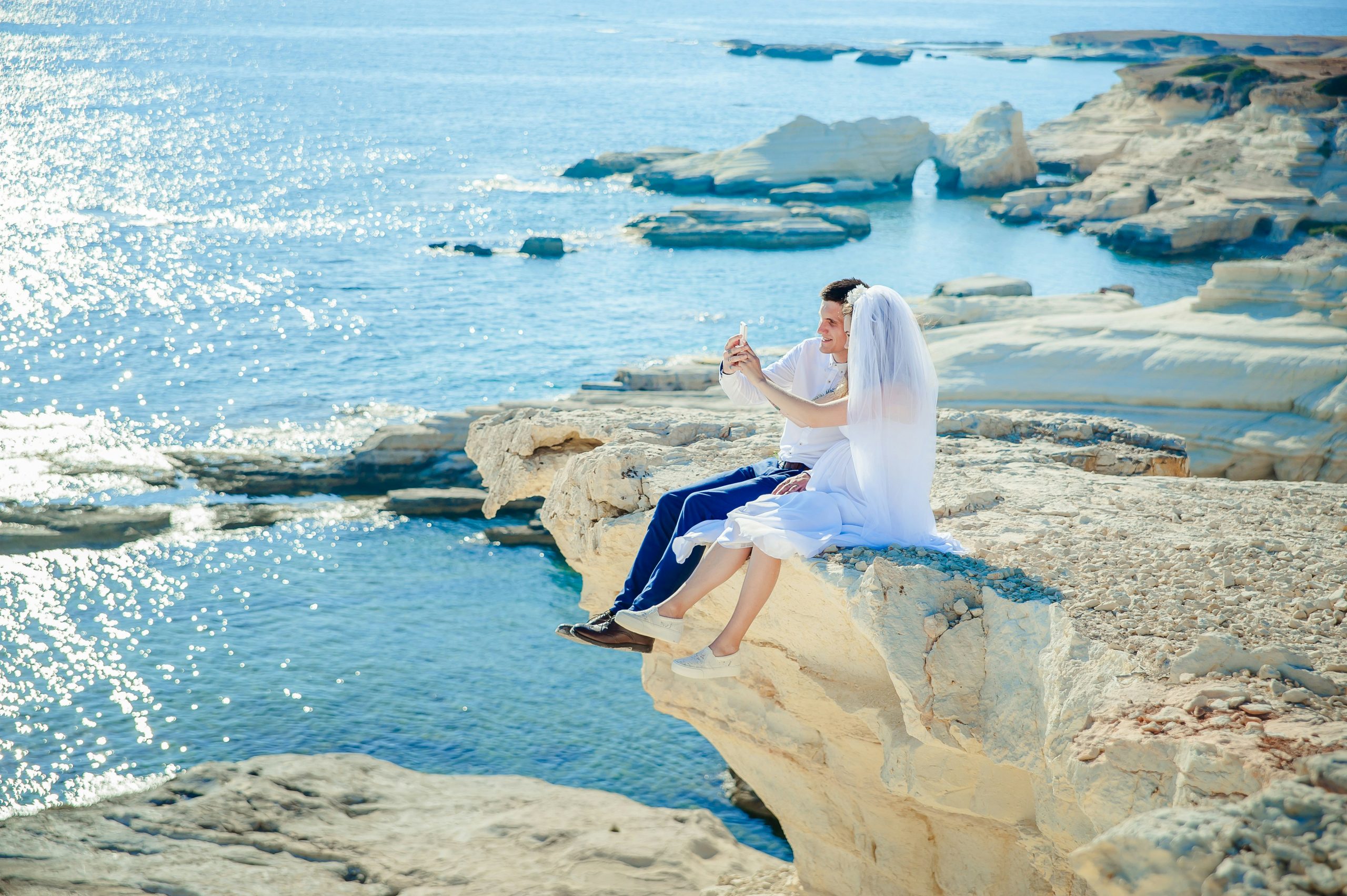 A couple sitting on a high rock overlooking the blue ocean, taking a photo together during their honeymoon