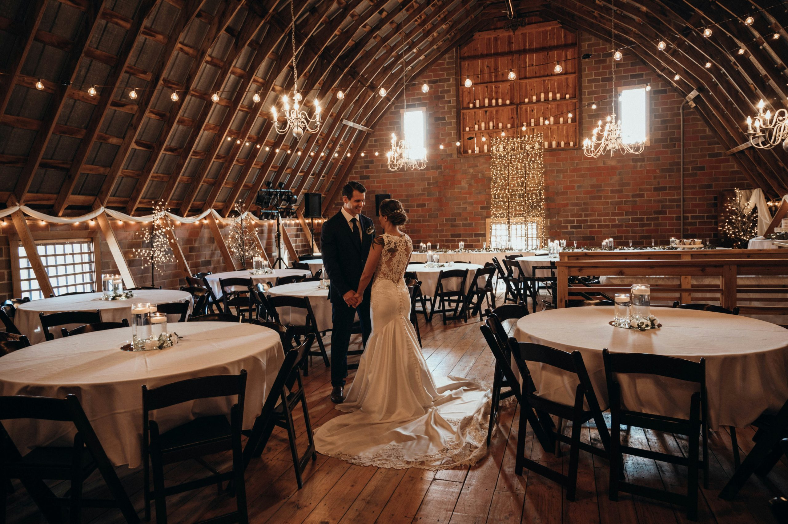 A couple standing in the center of a barn-style wedding venue, holding hands and looking at each other in an intimate moment