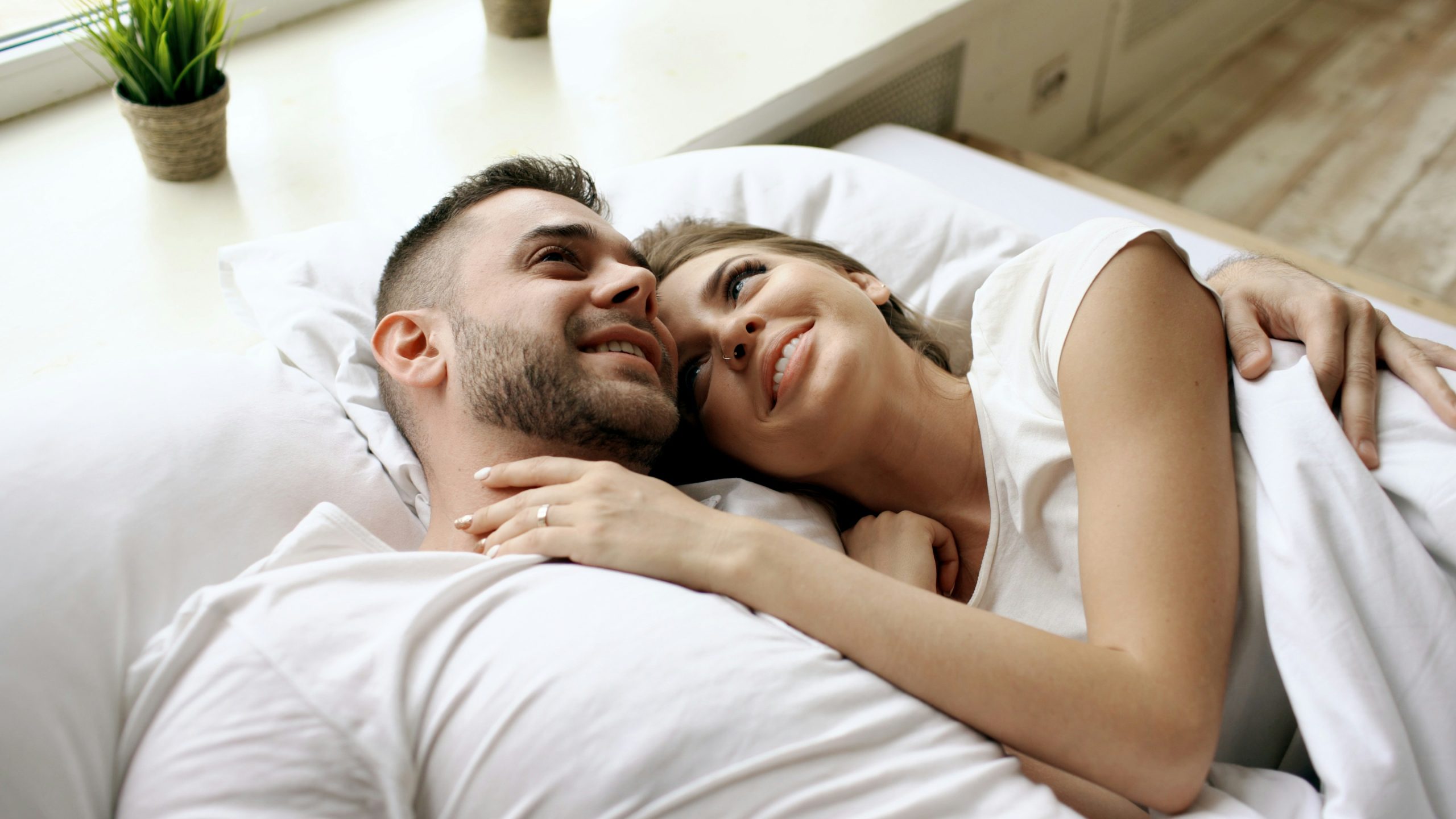 A couple lying down together, with the woman resting on the man’s shoulder as they smile and look into each other’s eyes during a relaxed moment