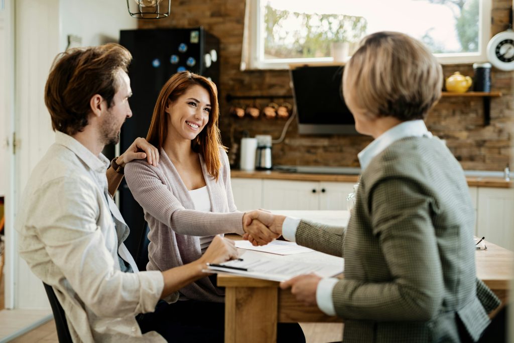 A couple standing in a house, with the man’s hand on his partner’s shoulder as she shakes hands and smiles at a real estate agent holding documents during a home buying process