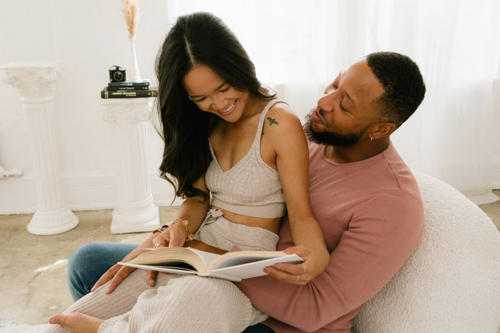 A happy couple sitting on a beanbag, with the woman on the man’s lap as they look at a book and laugh together in a relaxed moment