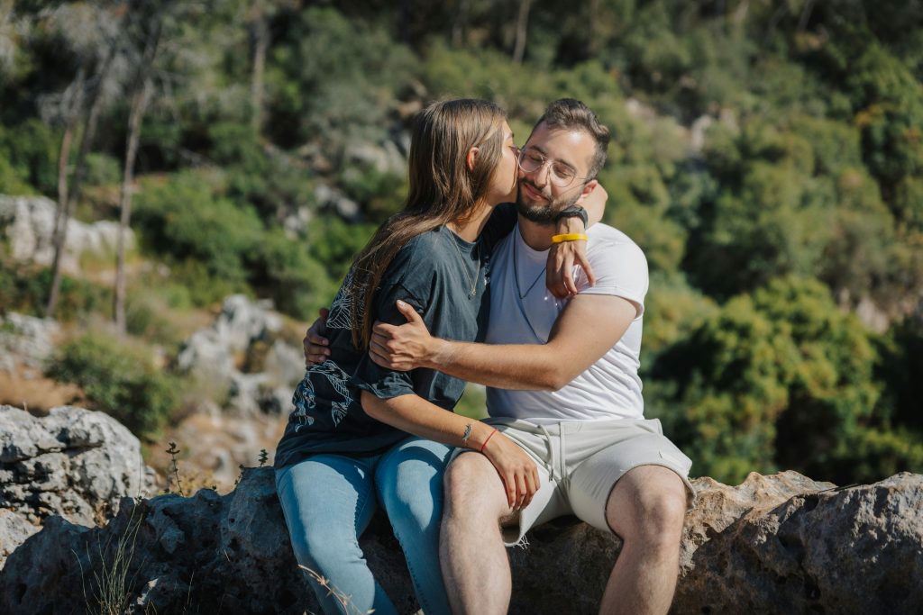 A couple sitting on a rock with a natural landscape in the background, as the woman kisses the man on the cheek while he closes his eyes