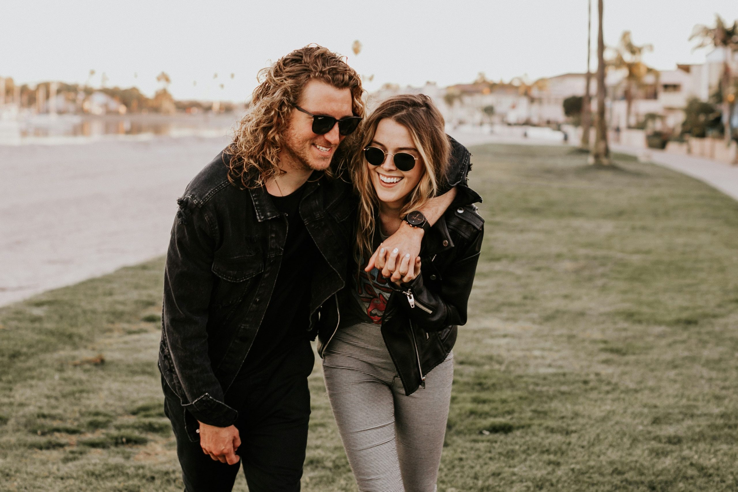 A smiling couple wearing jackets and sunglasses, walking arm in arm along the beach on a chilly day
