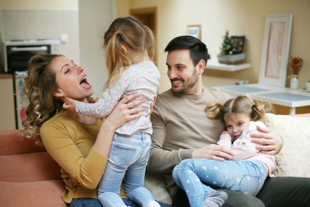 A joyful family with two young girls playing on a sofa, all laughing and enjoying a happy moment together