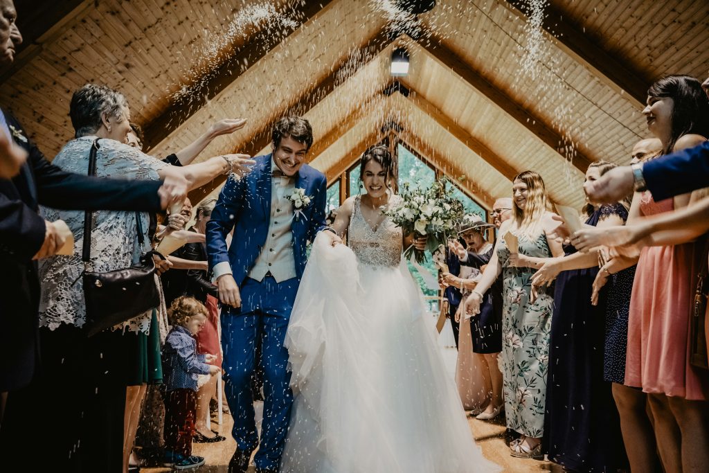 A happy couple walking under a wooden-roof venue as guests throw rice over them, celebrating their wedding exit with joy