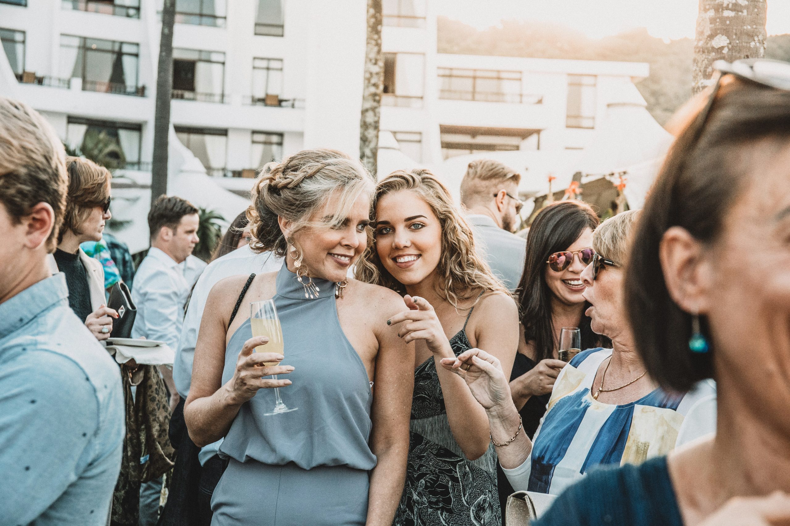 Two women posing at a wedding celebration Two women smiling and posing for the camera at a wedding, with other guests visible in the background during the celebration