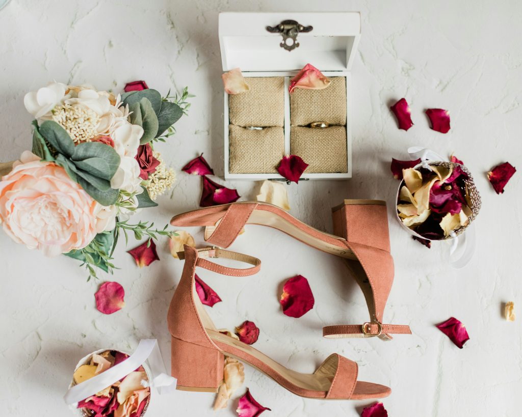 A styled arrangement of wedding shoes, a bridal bouquet, and wedding rings surrounded by flower petals for a romantic detail shot