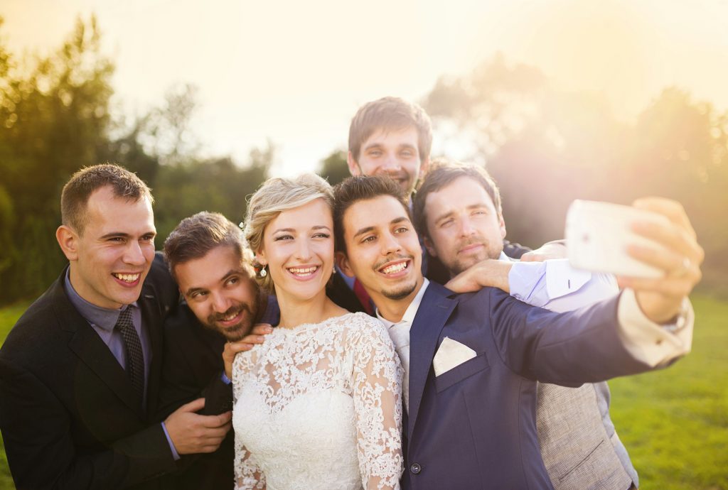 A group selfie at a wedding featuring the newlyweds, their family, and friends smiling together during the celebration