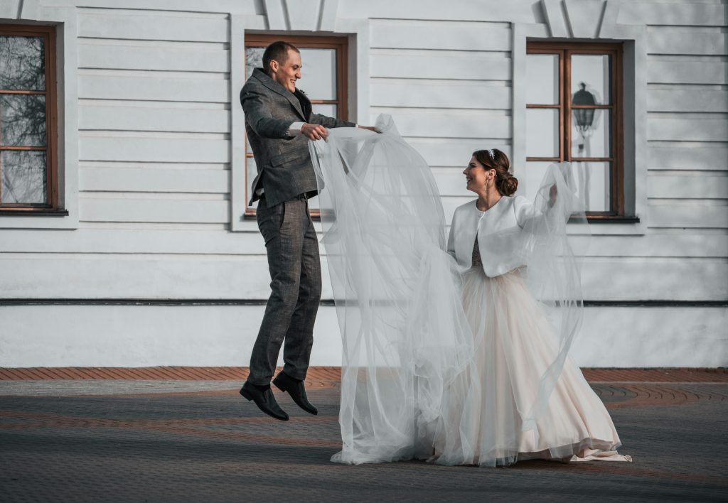 A groom playfully lifting or adjusting the bride’s dress as they laugh together, sharing a joyful moment on their wedding day