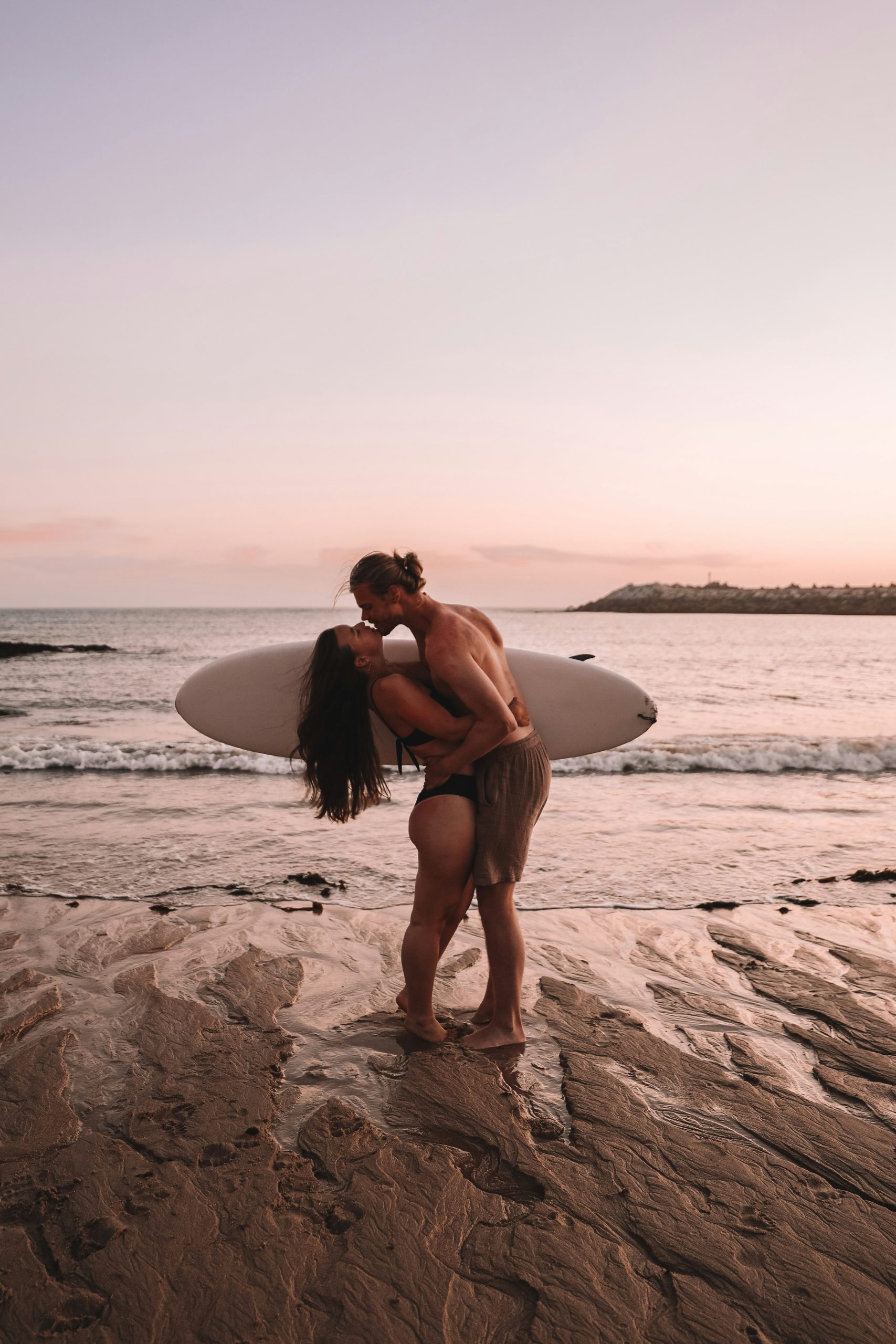 California couple kissing at sunset on the beach with a surfboard