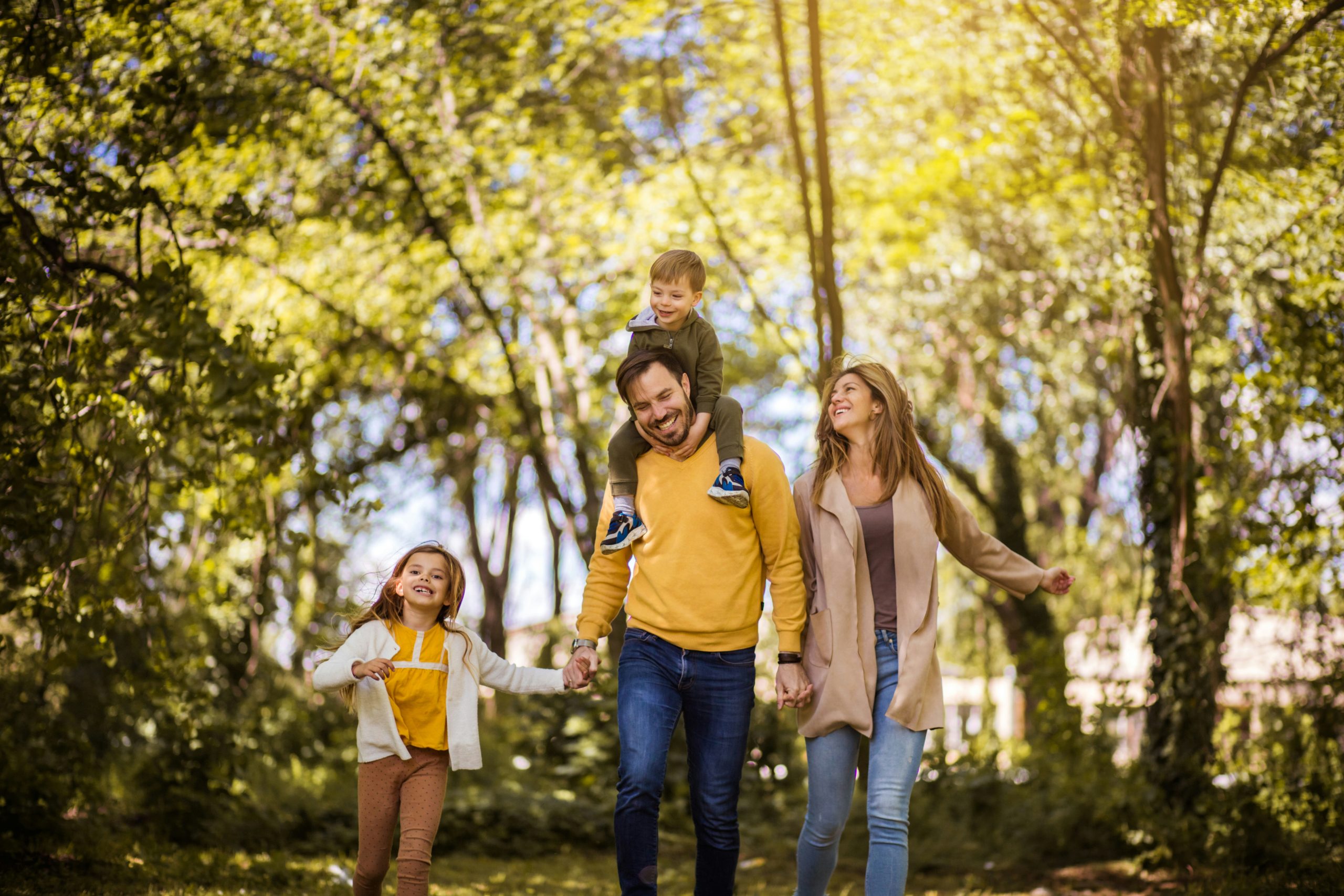 A happy family with two children walking through a park on a sunny day, with one child riding on the father's shoulders while everyone laughs and enjoys time together outdoors.