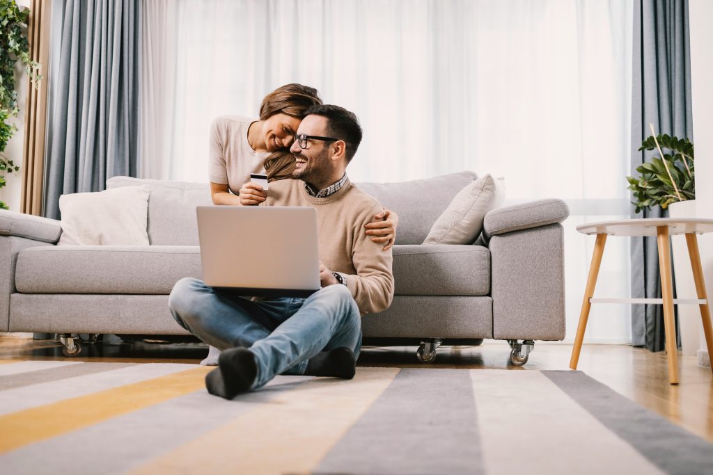 A happy couple looking at a smartphone and laptop while holding a payment card, making an online purchase together and smiling.