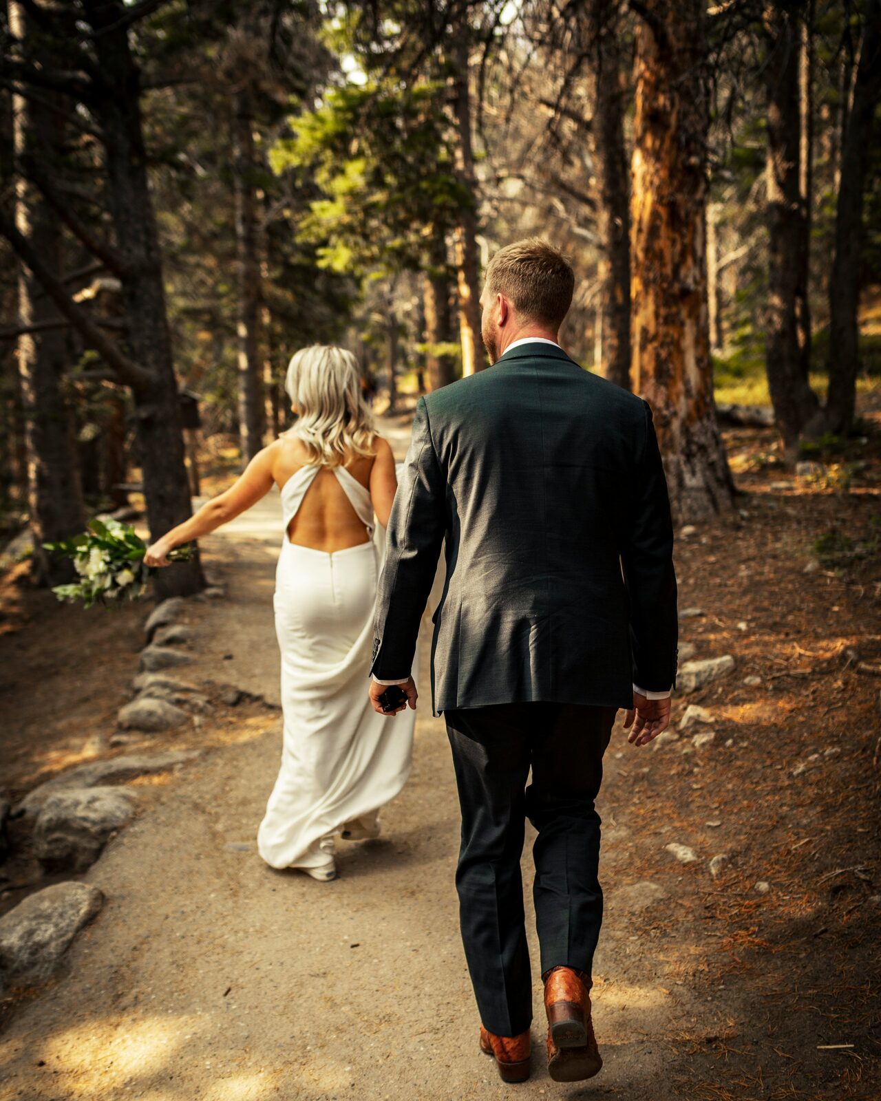 Colorado bride and groom walking a forest path with a ring box, seen from behind