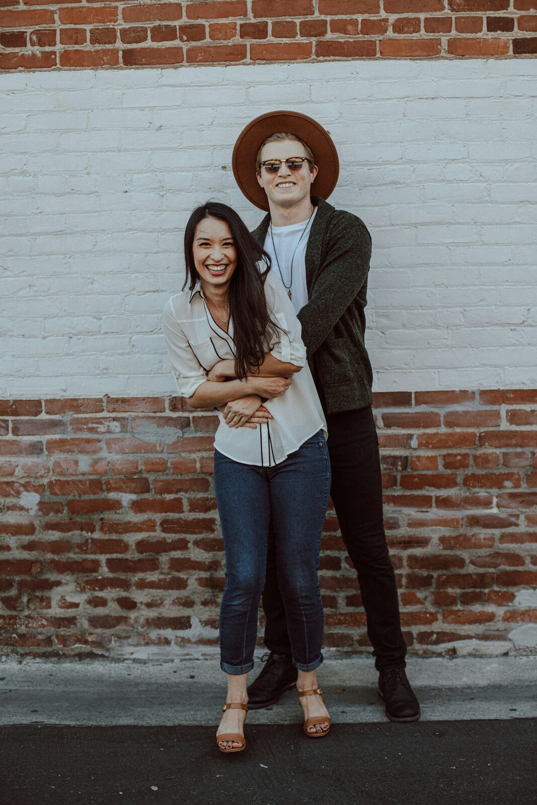 Virginia couple in casual engagement style against a brick wall, one in a wide-brim hat
