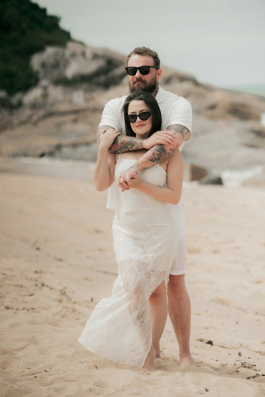 Tattooed Virginia groom embracing his bride in a white lace dress on a sandy beach