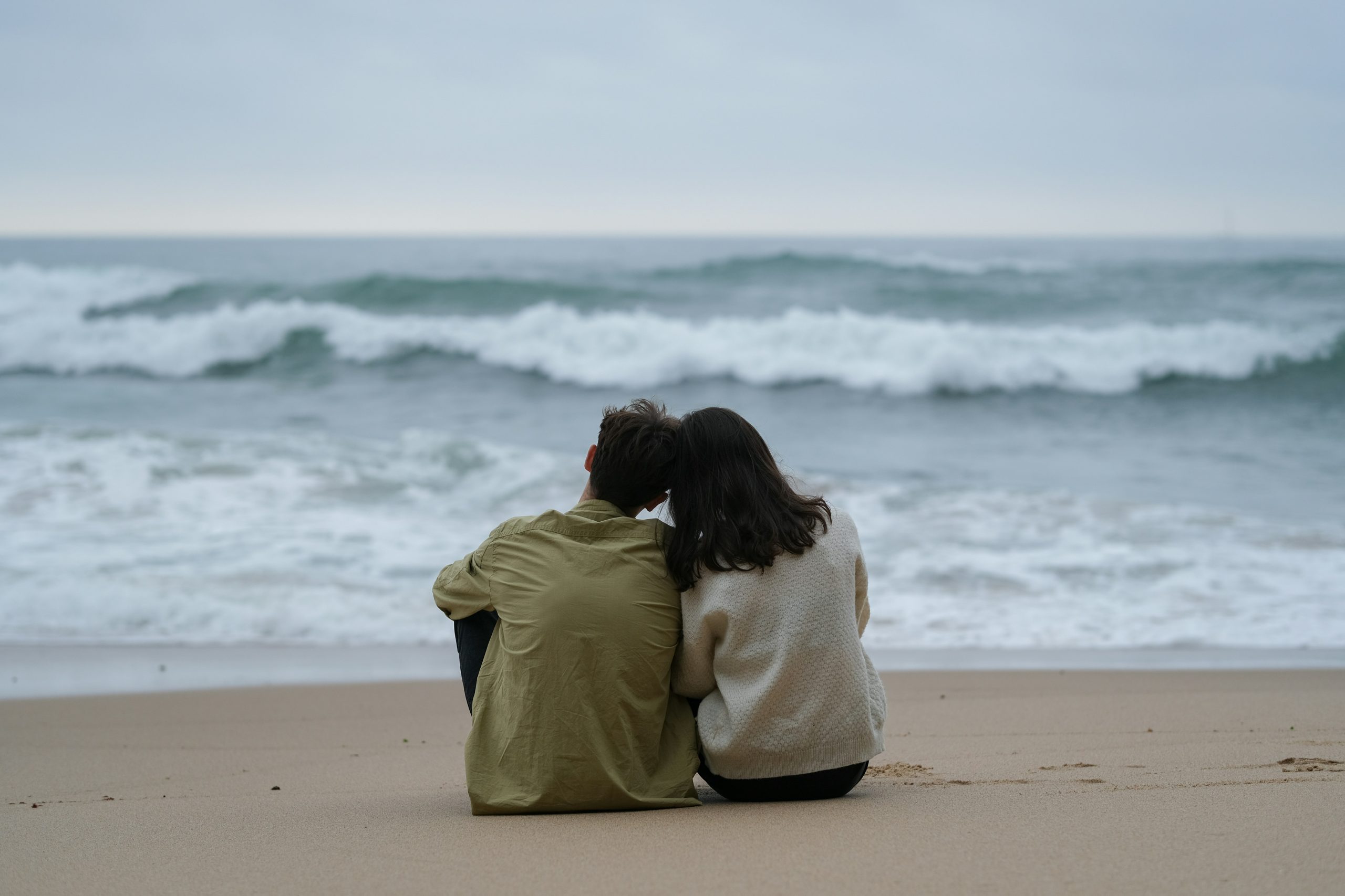 Couple sitting together by the ocean A couple sitting on the sand and looking out at the ocean, with their heads gently resting against each other in a peaceful moment.