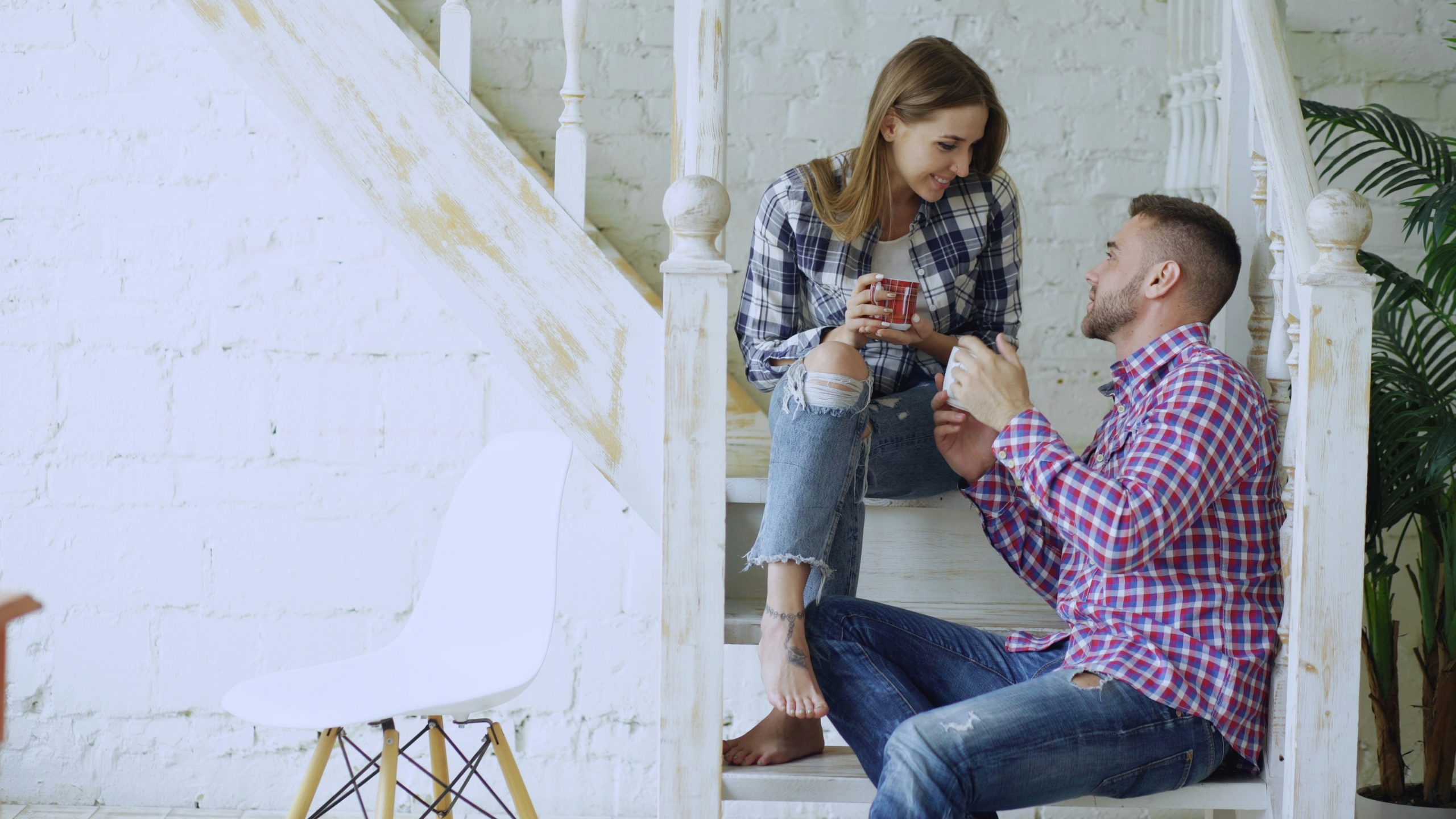 A couple sitting on the stairs, talking and smiling while enjoying warm drinks together in a cozy setting.