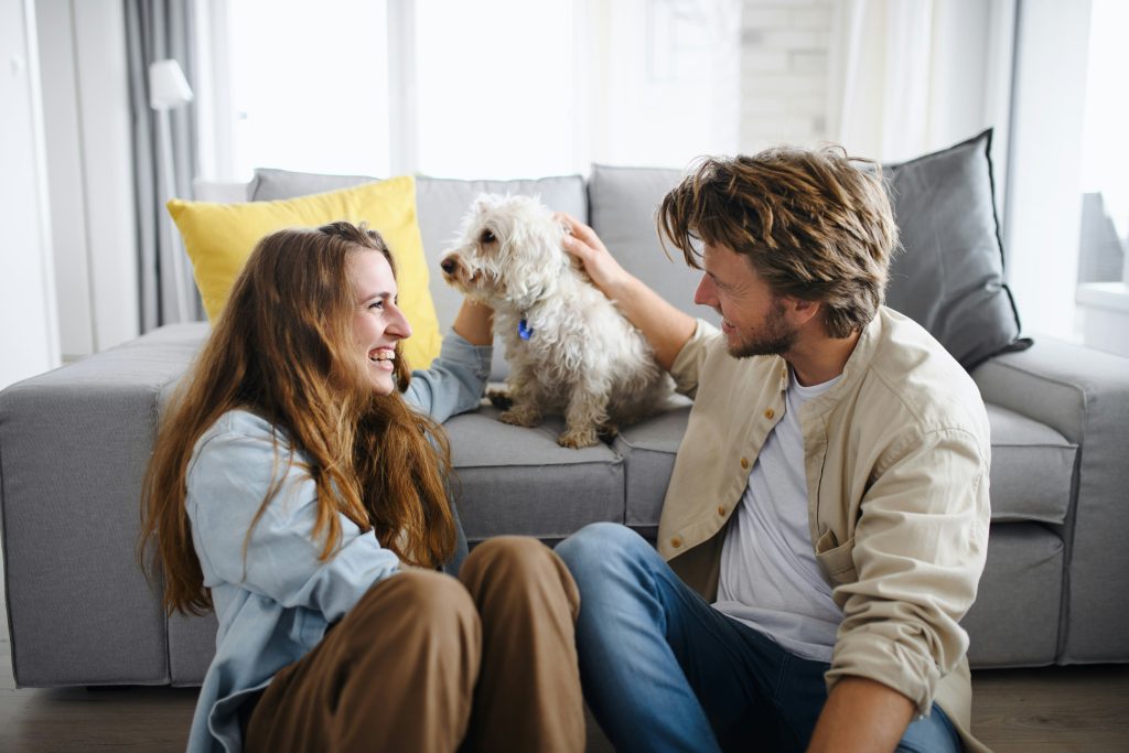 A couple sitting on the floor in front of a sofa, laughing and talking while gently petting their dog together.