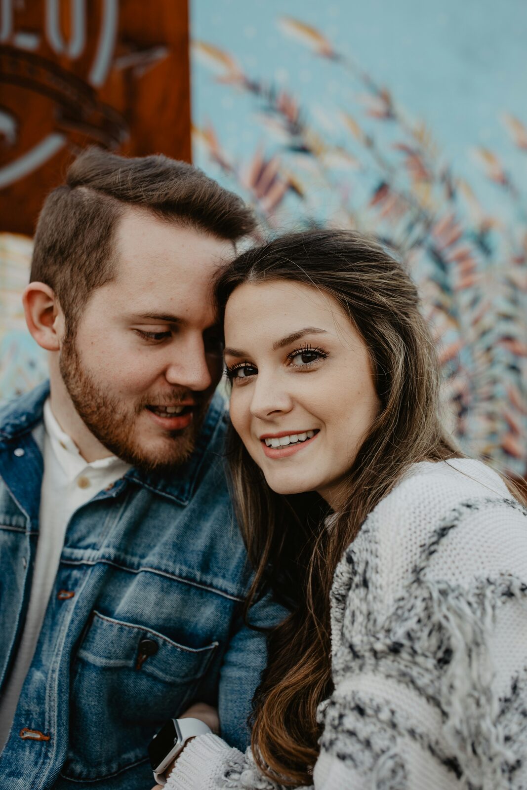 Close-up engagement portrait of a Connecticut couple by a mural wall, in a denim jacket and fluffy cardigan