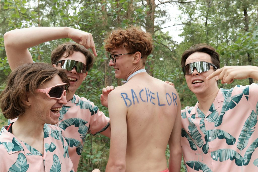 A groom with the word “Bachelor” on the back of his outfit poses with his friends, who wear funny sunglasses during a lively bachelor party.