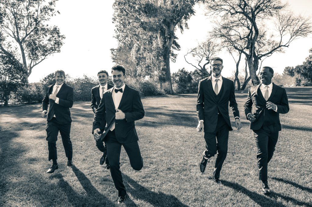 A groom running and laughing with his friends, all dressed in formal wedding attire, during a fun moment before the ceremony.