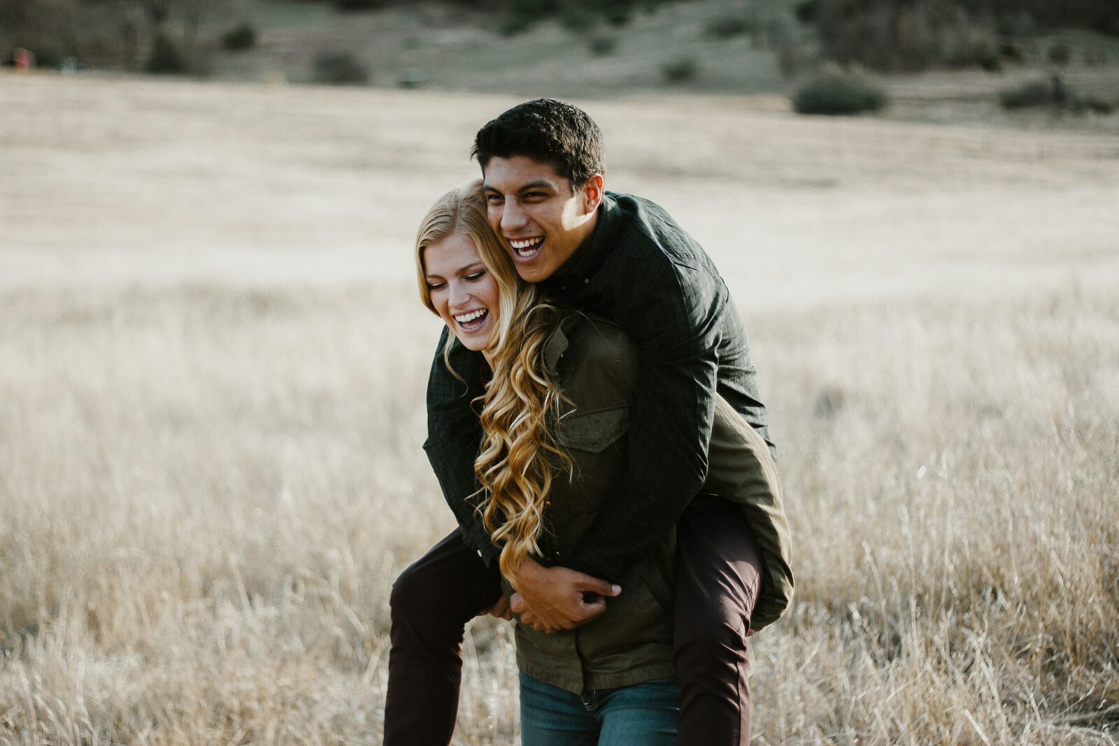 Engaged couple laughing during a piggyback ride in a golden grass field, thinking about the property they will share