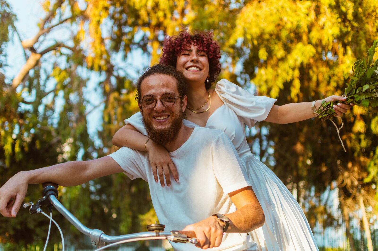 Just-married Louisiana couple riding a bike together under willow trees, bride holding a eucalyptus bouquet