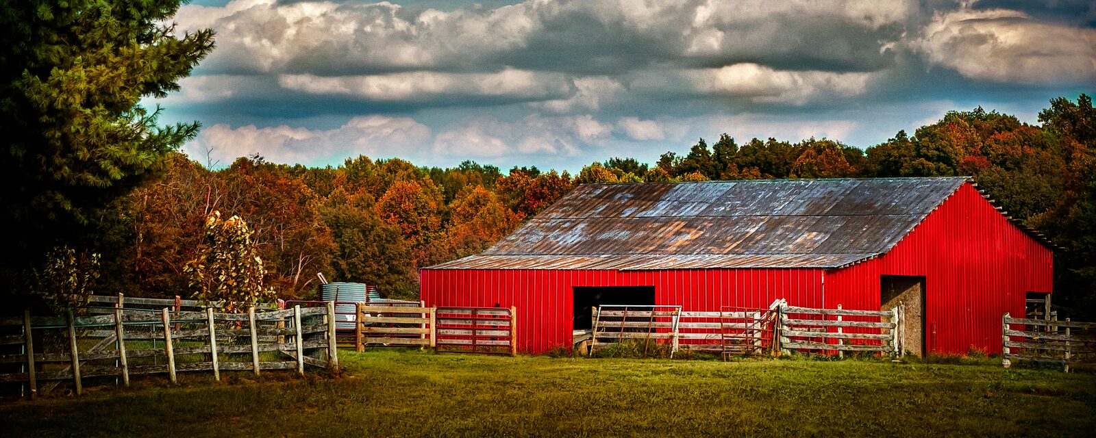 Red barn with a tin roof behind a wooden fence, surrounded by autumn foliage in the countryside